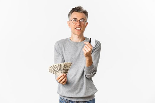 Portrait of excited middle-aged man looking amazed, holding money and showing credit card, standing over white background