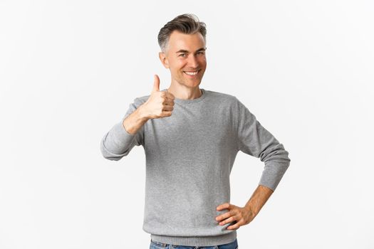 Portrait of proud and happy middle-aged man, smiling pleased and showing thumbs-up, praise something good, standing over white background