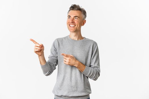 Image of smiling handsome middle-aged man looking at logo, pointing fingers left and showing advertisement, standing over white background