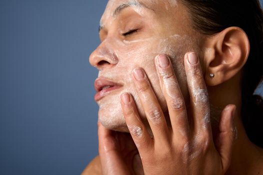 Head shot of beautiful woman with perfectly healthy skin, holding hands on her face, performing smoothing massage, removing makeup and cleansing face with exfoliating scrub and facial foam cleanser