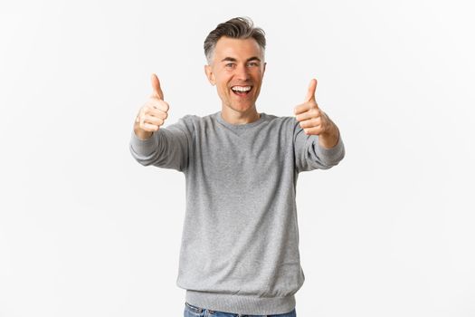 Portrait of cheerful middle-aged man praising good work, standing over white background, showing thumbs-up in approval and smiling glad