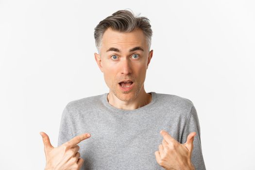 Close-up of confused middle-aged man, pointing at himself and gasping, standing over white background