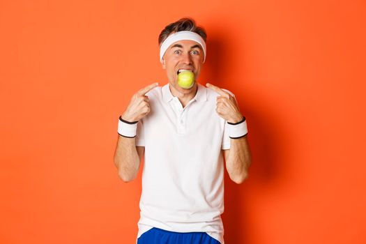 Portrait of funny, handsome middle-aged fitness guy, holding apple with teeth and pointing at it, eating healthy and working out, standing over orange background