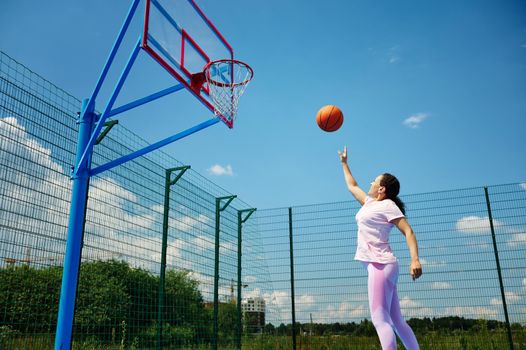 Young woman playing basketball on the court outdoor and throwing the ball into the hoop
