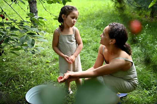 Adorable girl with her mother eating cherries while picking in the garden. Happy motherhood and childhood, love, tenderness. Enjoying time together. Happy family. Positive human emotions.