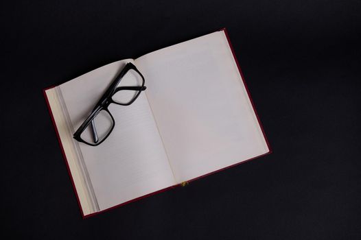 Flat lay of eyeglasses on an opened book in hard red cover, isolated over black background with space for text. Teacher's Day concept, Knowledge, literature ,reading, erudition