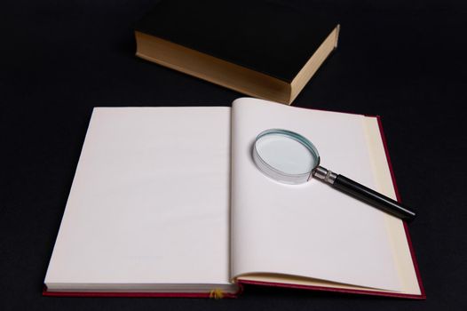 A magnifying glass, loupe, magnifier on an opened book in hard red cover, isolated over black background with space for text. Teacher's Day concept, Knowledge, literature ,reading, erudition