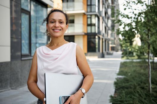 Happy brunette mixed race woman in casual attire holding laptop computer, smiling looking at camera on the tall buildings background. Business, freelance, office work concept