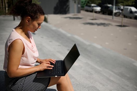 Side portrait of busy freelance woman, start-uper , office worker using laptop sitting on steps during coffee break. Office worker on lunch break in urban background