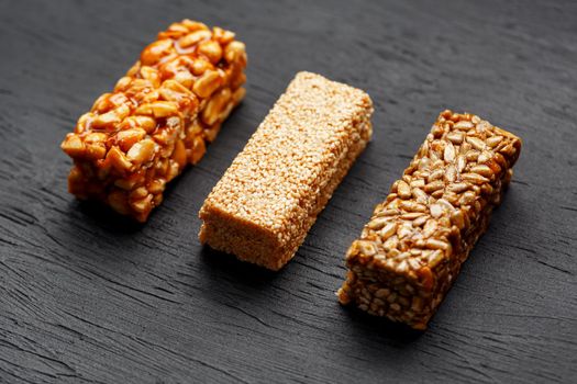 Cereal granola bar with peanuts, sesame and sunflower seeds on a cutting board on a dark stone table. View from above. Three Assorted Bars