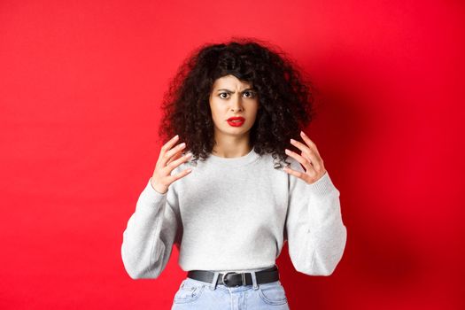 Angry caucasian woman frowning and raising hands mad, wants to strangle or kill someone annoying, standing on red background