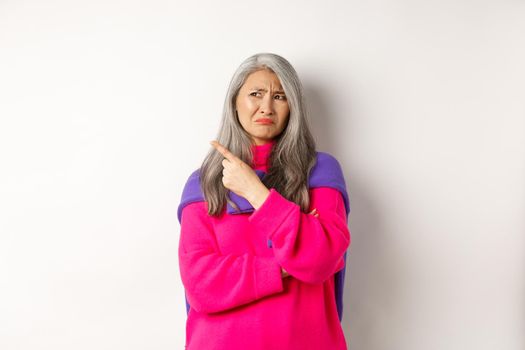 Disappointed asian grandmother grimacing disgusted, pointing finger left and complaining on something bad, standing over white background
