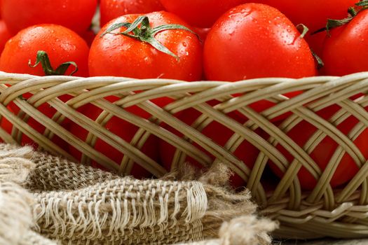 Ripe and juicy cherry tomatoes with drops of moisture in a wicker basket. Old wooden table, around the canvas of burlap