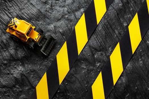 Yellow toy bulldozer on a black background texture. Black and yellow fence tape against the background of the CONCEPT