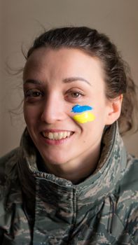 Indoor portrait of young girl with blue and yellow ukrainian flag on her cheek wearing military uniform, mandatory conscription in Ukraine, equality concepts