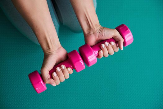 Woman hands holding pink dumbbells on green sport mat background. Home exercise during pandemic. Workout in gym. Fitness concept.