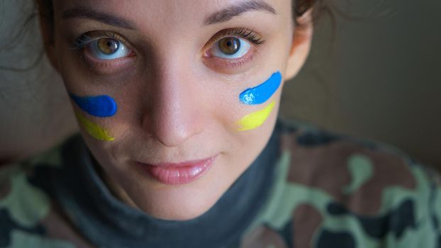 Indoor portrait of young girl with blue and yellow ukrainian flag on her cheek wearing military uniform, mandatory conscription in Ukraine, equality concepts