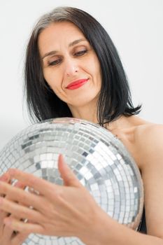 Studio photo of middle aged woman starting getting grey-haired wearing black clothes with silver disco ball in hands on white background, middle age sexy lady, happy life concept