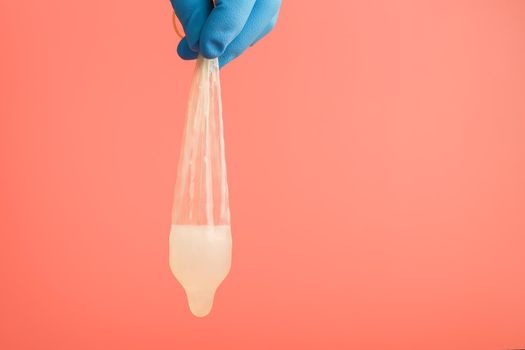 A doctor s hand in a blue glove holds a used condom with sperm on a pink background. Sperm latex and protection against pregnancy.