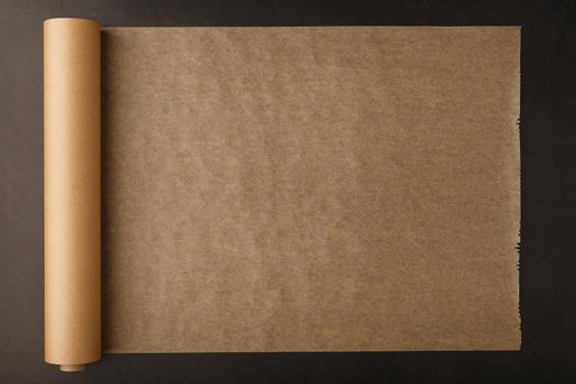 A roll of unfolded brown parchment paper, for baking food in on a dark background, top view.