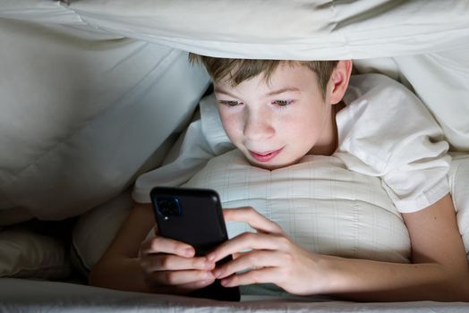 Boy under the blanket at night in his bed communicates on Internet. Child gadget addiction and insomnia. social media addiction