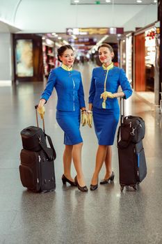 Full length of women stewardesses in aviation air hostess uniform looking at camera and smiling while standing near trolley luggage bags