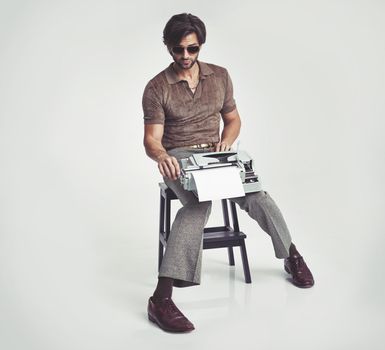 Who needs a desk anyway. A handsome man holding a retro typewriter and smoking a pipe while sitting on a stool.