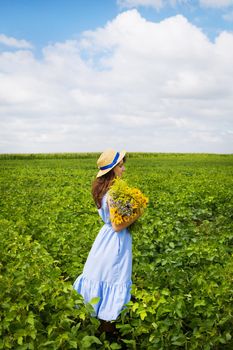 beautiful girl stands in the field with a bouquet of yellow flowers and a straw hat