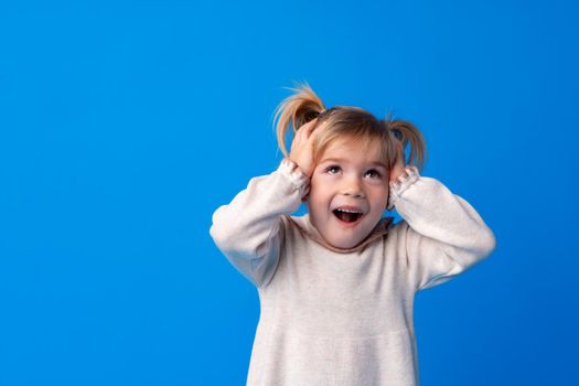 Beautiful little girl with hands on head being delighted and shocked over blue background.