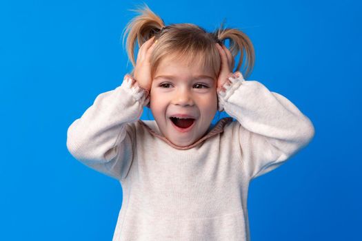 Beautiful little girl with hands on head being delighted and shocked over blue background.