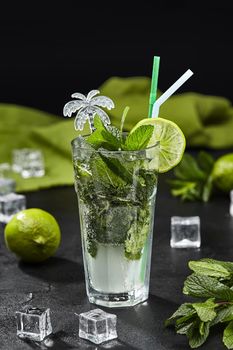 Glass of non-alcoholic mojito cocktail traditionally made with lime, fragrant mint and ice on table with ingredients against black background. Popular summer refreshing drink concept