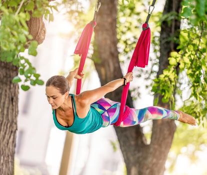 Beautiful girl during fly yoga in hammock at nature. Young sport woman doing aero fitness gymnastic stretching outdoors