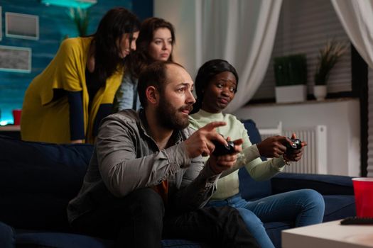 Excited multiethnic friends sitting on couch holding controller playing videogames