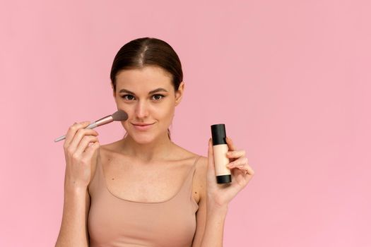 A beautiful model applies foundation with a brush to clean skin on pink background