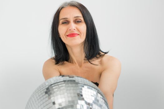 Studio photo of middle aged woman starting getting grey-haired wearing black clothes with silver disco ball in hands on white background, middle age sexy lady, happy life concept