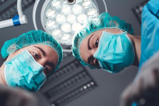 From below female surgeons in medical uniform using professional tools while standing under bright light in operating theater
