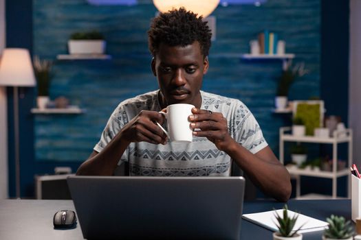 Businessman holding cup drinking coffee while reading business project using laptop computer working remote from home. Freelancer man sitting at desk during online lesson in living room