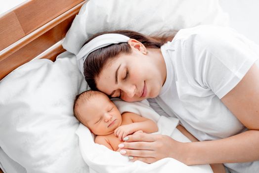 Beautiful smiling mother lying in the bed with sleeping newborn baby and napping. Young girl resting with infant child at home. Mom with kid together at home
