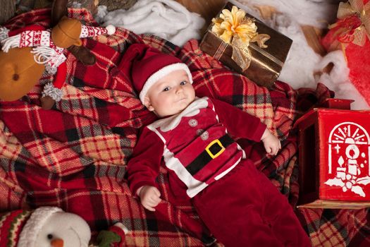 A child in Santa Claus costume rests on a red plaid surrounded by toys.