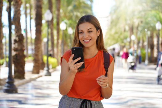 Hispanic young woman laughing and texting on her mobile phone walking outdoors