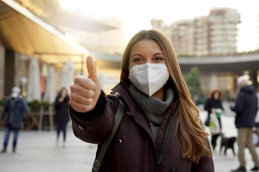 Thumbs up woman wearing protective face mask and winter clothes in the city