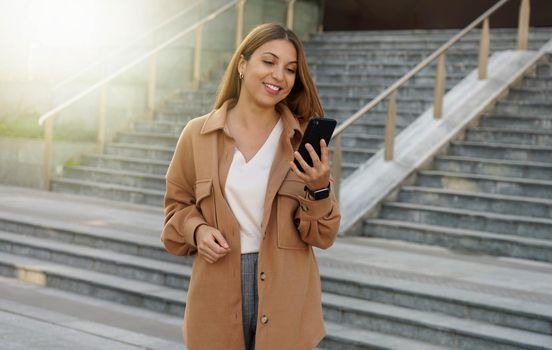 Portrait of successful businesswoman holding mobile phone while walking outdoors near financial office, young woman entrepreneur using smartphone