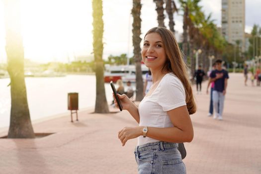 Teenager girl using her cellphone on the waterfront while strolling at sunset