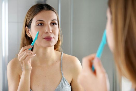 Beautiful young woman shaving her face by razor at home. Pretty woman using razor on bathroom.