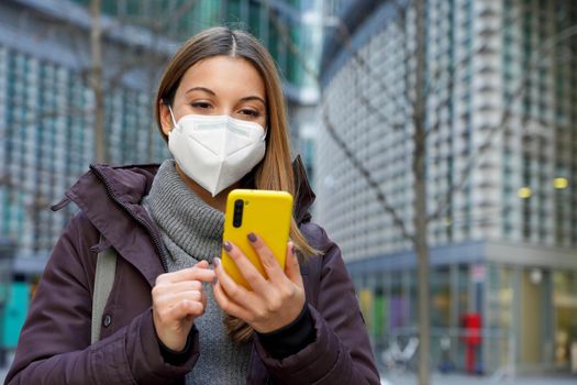 Portrait of a young woman with medical face mask checking notifications on smartphone on urban background. Copy space.