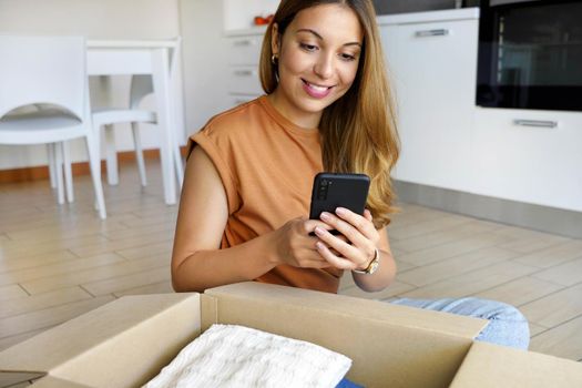 Online store selling clothes on website working on smartphone ecommerce business from home. Woman packing clothing purchase in shipping packages for delivery.