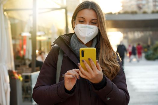 Portrait of a young woman with medical face mask walking in the city using smartphone on sunset