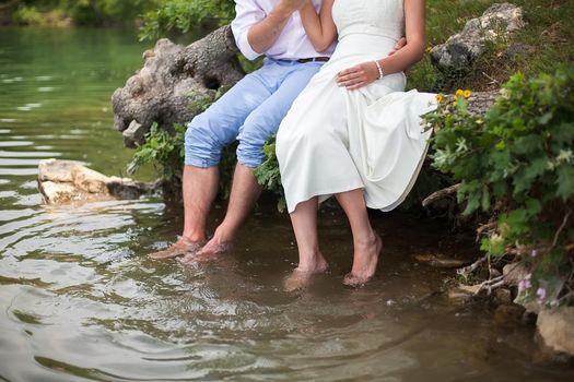 The bride and groom sitting on the shore of the lake. Foot splashing in the water