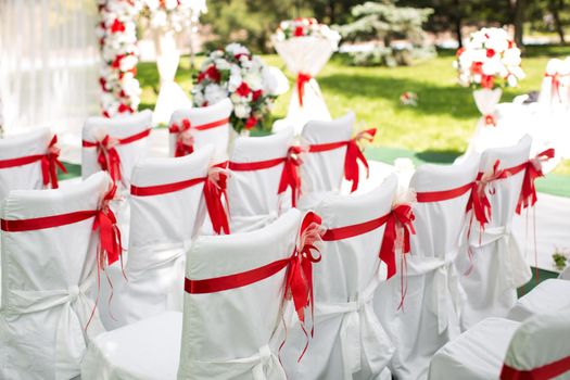 Wedding ceremony outdoors. White chairs with red ribbon