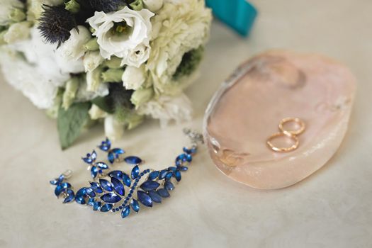 Gold wedding rings on a pink marble shell next to blue jewelry and flowers.